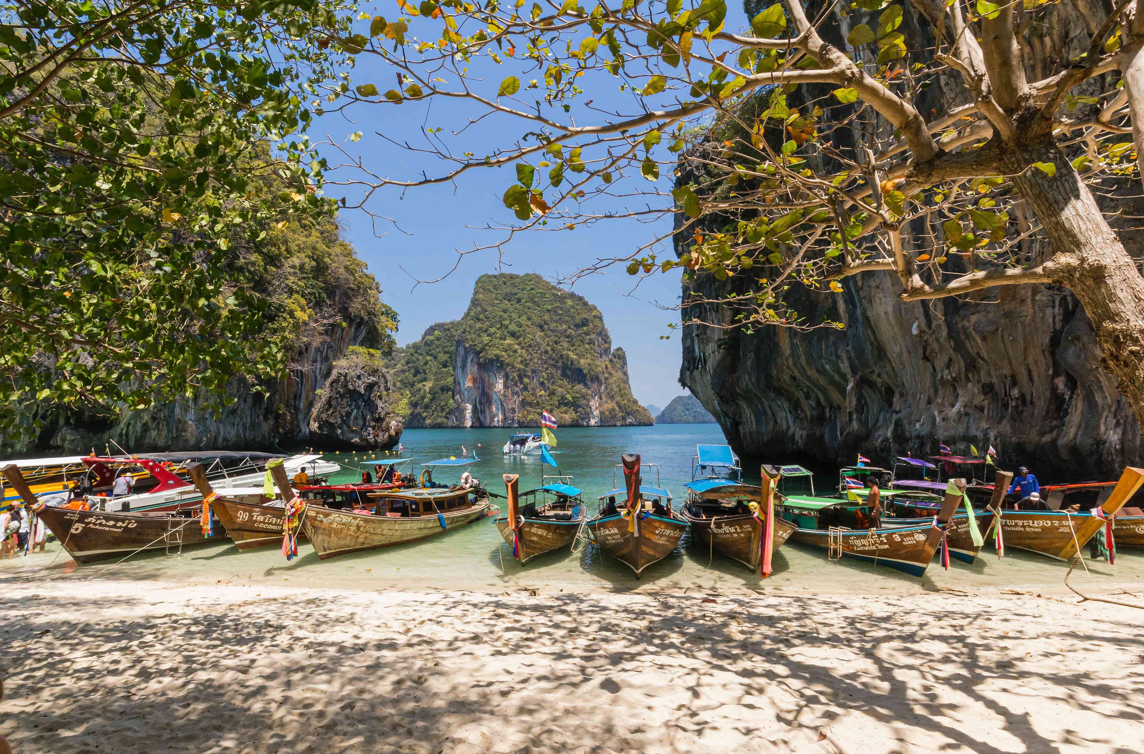 A picturesque beach scene in Thailand, where traditional long-tail boats are gently moored on the soft, golden sand. The boats, with their colorful wooden hulls and long, decorative prows, are adorned with vibrant ribbons and flowers, swaying lightly in the breeze.