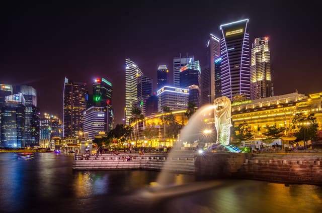 A majestic, half-lion, half-fish statue, known as the Merlion, stands proudly overlooking Singapore's Marina Bay. The Merlion's head, with its piercing eyes and open mouth, spouts a steady stream of water into the bay. Its fish body, sleek and powerful, rests on a concrete base. The statue is set against a backdrop of modern skyscrapers and lush greenery, creating a stunning contrast between the old and the new. The park surrounding the Merlion is filled with visitors taking photos, enjoying the scenic views, and simply soaking up the atmosphere of this iconic Singapore landmark.